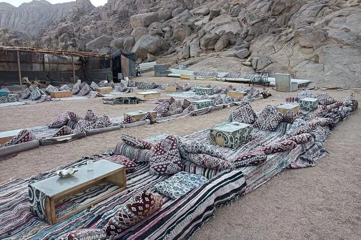 Une vaste salle à manger bédouine extérieure avec des rangées de tapis à motifs, des coussins et des tables basses entourée de montagnes à Charm el-Cheikh