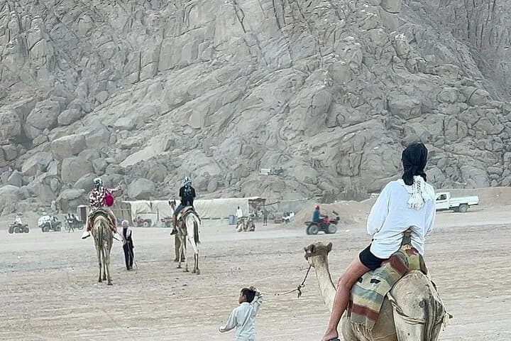 Des visiteurs à dos de chameau traversent une vallée désertique rocheuse avec des chameaux, des quads et des montagnes escarpées en arrière-plan à Charm el-Cheikh