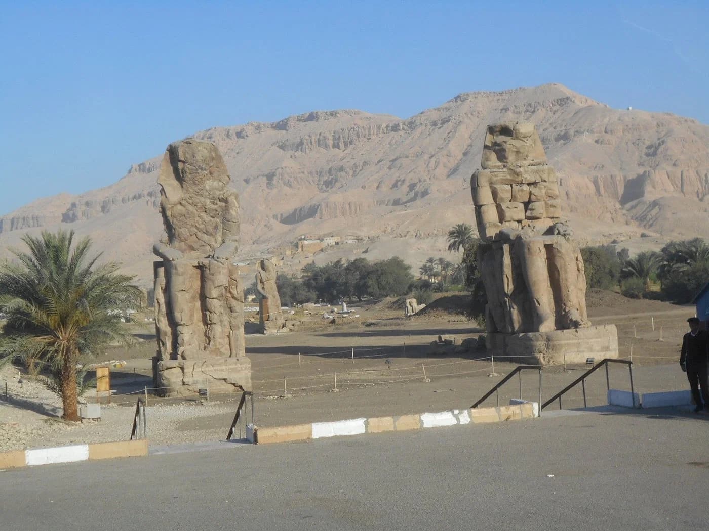 Two weathered Colossi of Memnon stand beside a road with palm trees and the Theban hills behind them in Luxor.