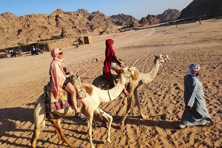 Deux visiteurs traversent à dos de chameau le camp de sable du désert tandis qu'un guide les accompagne à Charm el-Cheikh